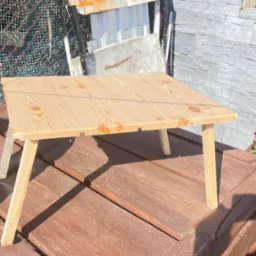 Surowy, sosnowy stolik kawowy z prostymi nogami, ustawiony na drewnianym tarasie, obok butelki wody i kwietnika z czerwonymi kwiatami, z widocznymi śladami obróbki drewna.