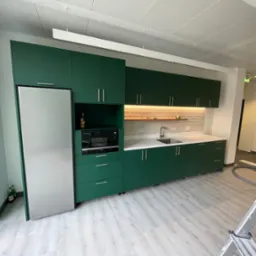 Nowoczesna kuchnia w biurze z zielonymi szafkami, białym blatem i sprzętem AGD, widoczny odkurzacz przemysłowy i drabina w tle.