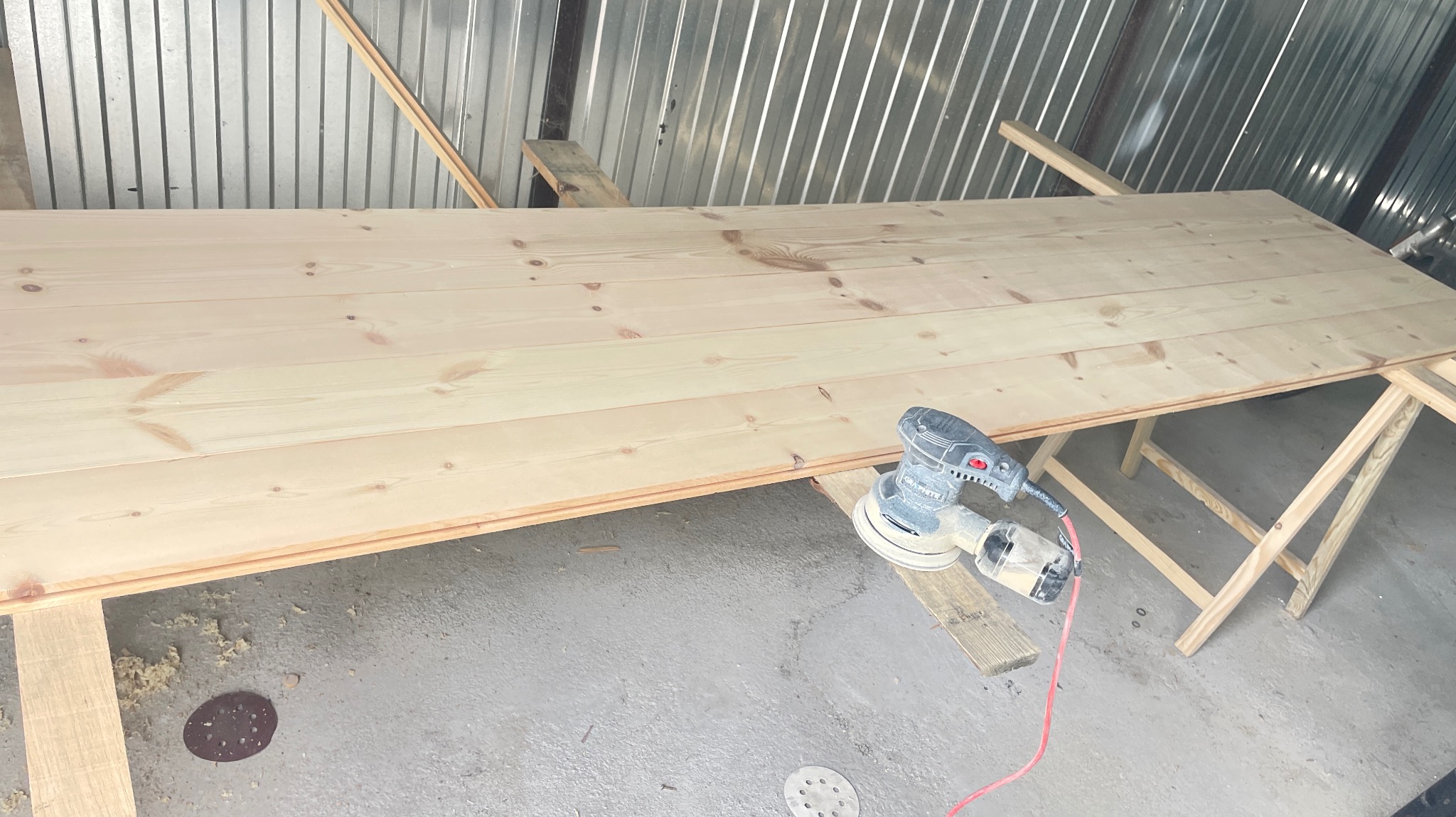 Duży, jasny blat z drewna sosnowego na kozłach, szlifowany szlifierką oscylacyjną, w tle blaszane ściany warsztatu.