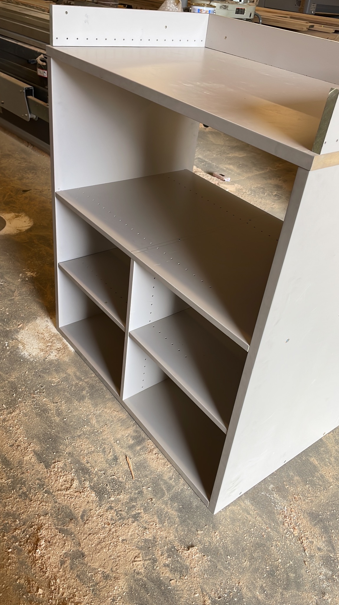 Surowy, biało-szary regał z ukośną górną półką i widocznymi otworami na półki, stojący na podłodze pokrytej wiórami.