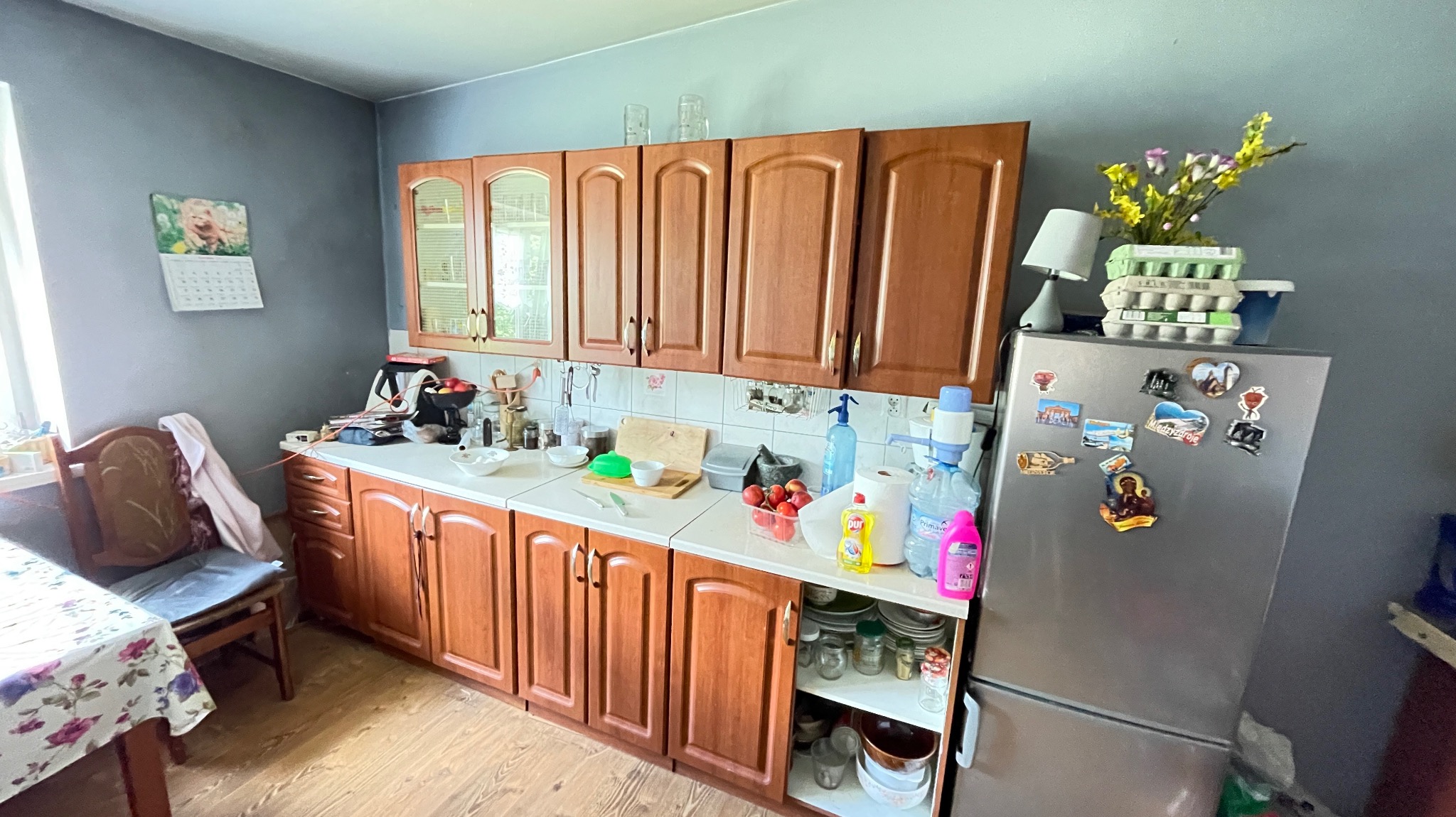 Kuchnia z drewnianymi szafkami, białym blatem, szarą ścianą i lodówką oblepioną magnesami; na blacie naczynia, owoce i środki czystości.