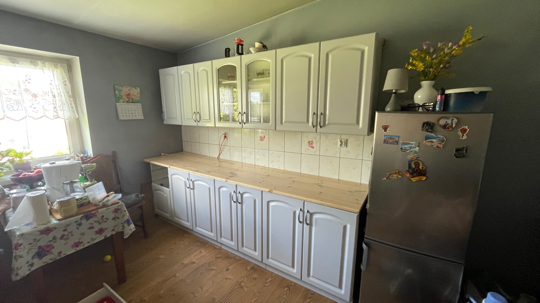 Szare szafki kuchenne z drewnianym blatem po renowacji w Trzcińsko-Zdroju, widoczne płytki ścienne z motywem kwiatowym i lodówka z magnesami.