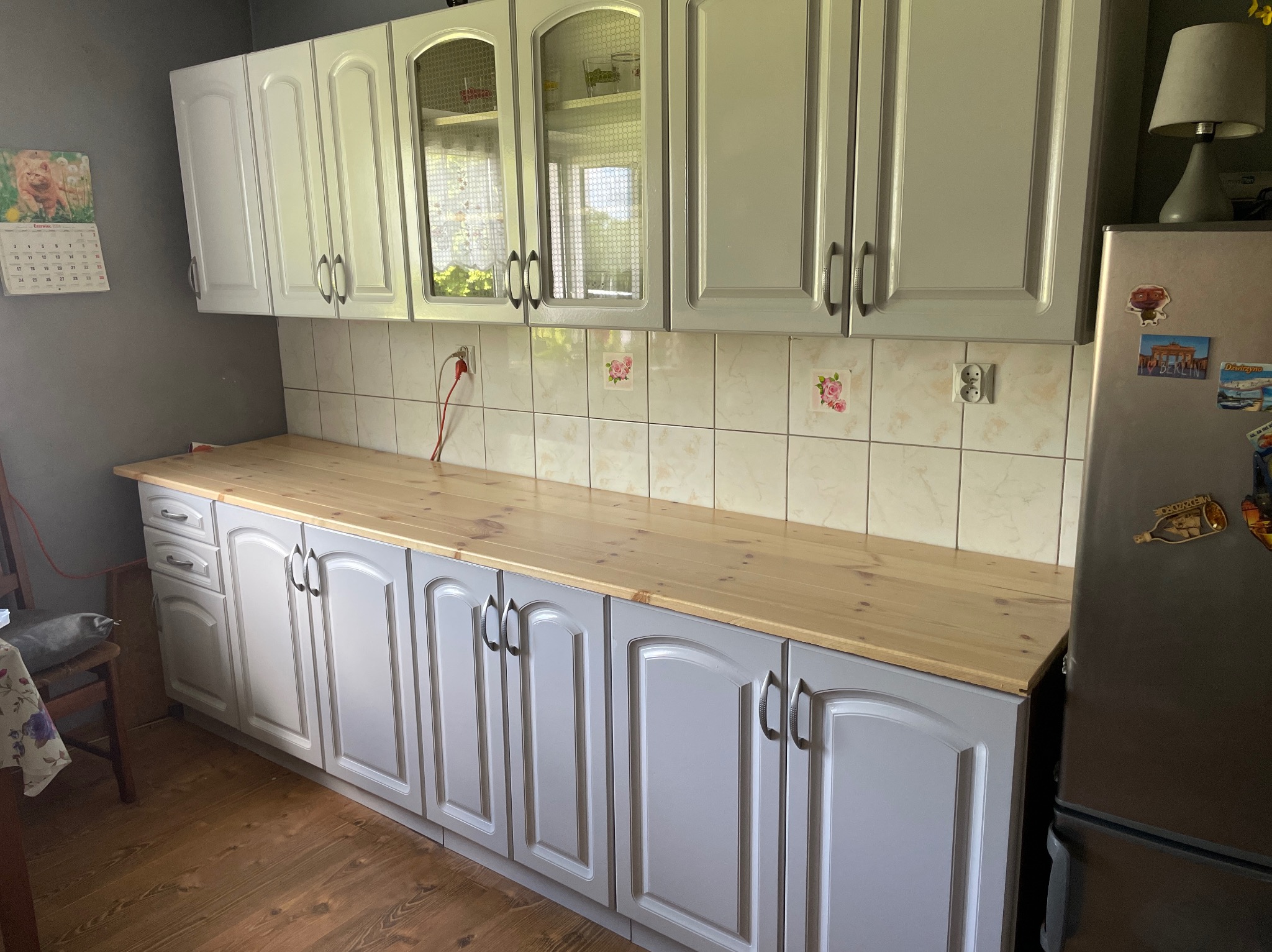 Szare szafki kuchenne z drewnianym blatem, widoczne płytki z motywem kwiatowym i lodówka z magnesami.