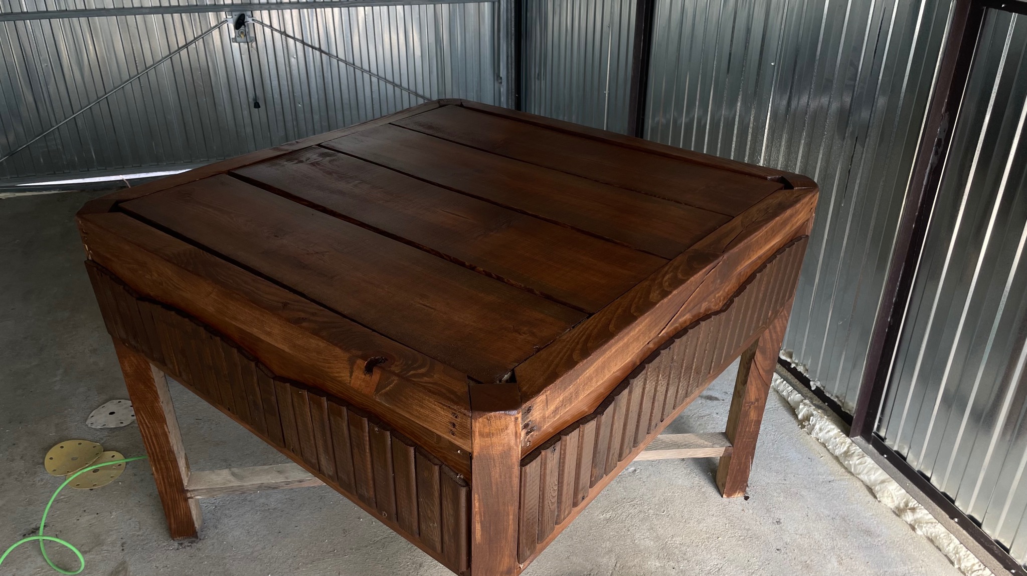 Ciemnobrązowy, kwadratowy stół z drewnianym blatem i rzeźbionymi bokami, stojący w garażu na tle blaszanych ścian, obok leżą krążki ścierne.