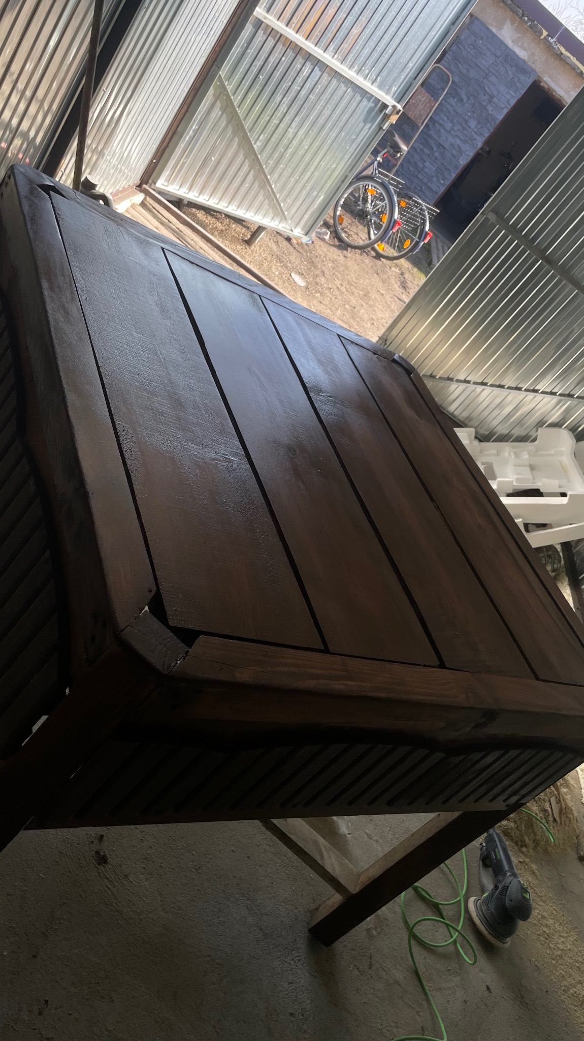Odnowiony drewniany stół ogrodowy z ciemnobrązowym blatem, widoczna szlifierka elektryczna na podłodze w tle, garaż blaszany i rower.