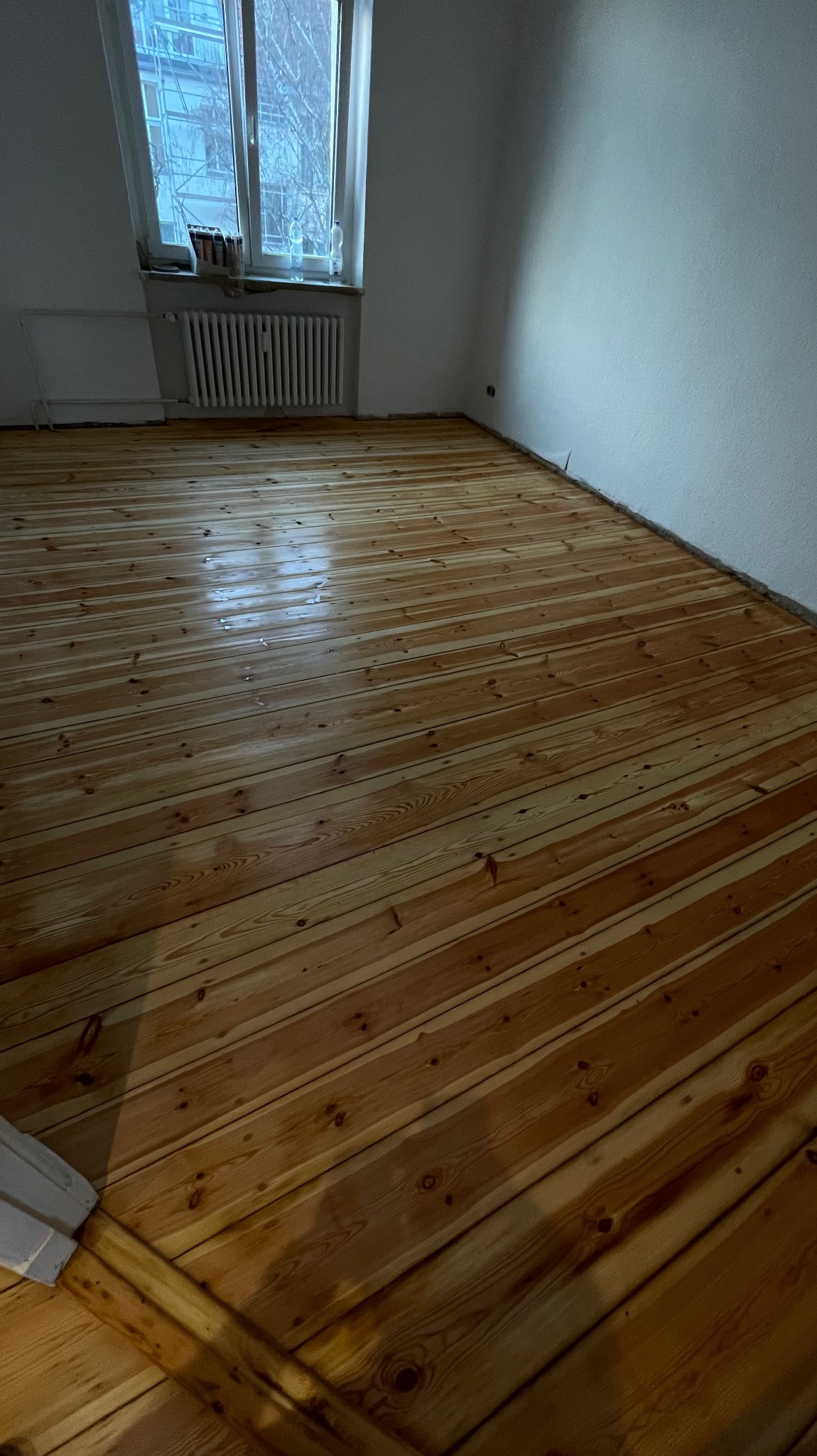 Świeżo wycyklinowana, lakierowana podłoga z desek w pustym pokoju z grzejnikiem pod oknem.