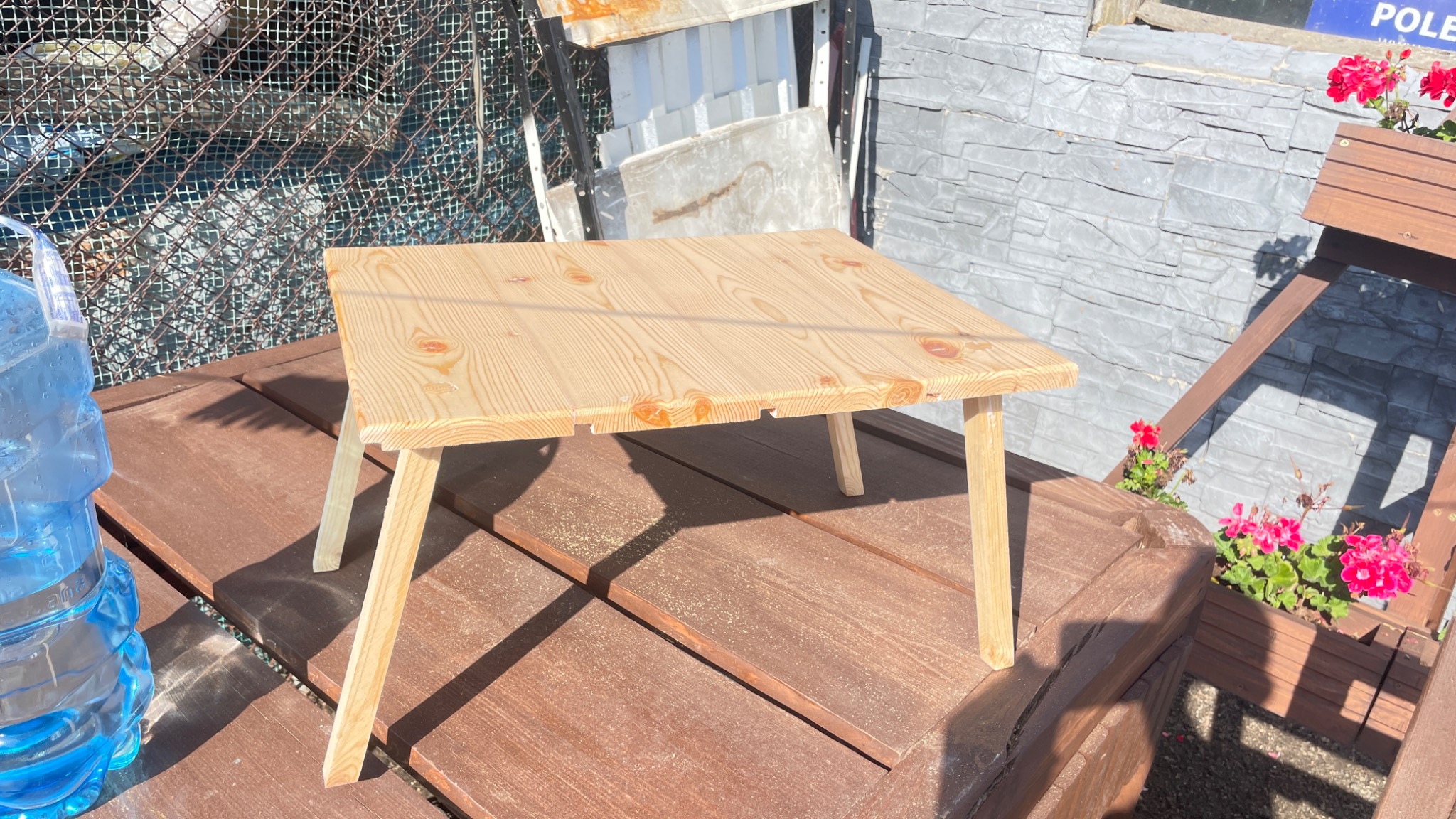 Surowy, sosnowy stolik kawowy z prostymi nogami, ustawiony na drewnianym tarasie, obok butelki wody i kwietnika z czerwonymi kwiatami, z widocznymi śladami obróbki drewna.