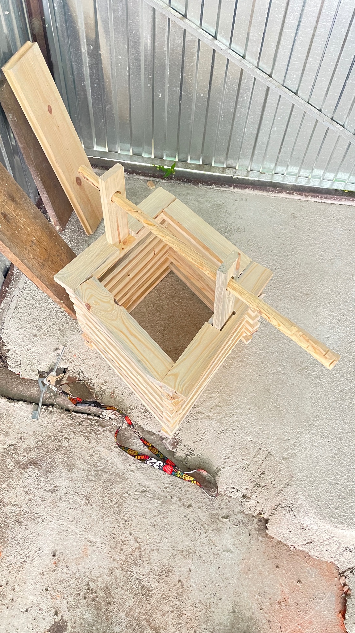 Drewniana konstrukcja studni z kantówki, bez wykończenia, z widocznym mechanizmem korbowym, leżąca na betonowej posadzce obok kluczy i smyczy.