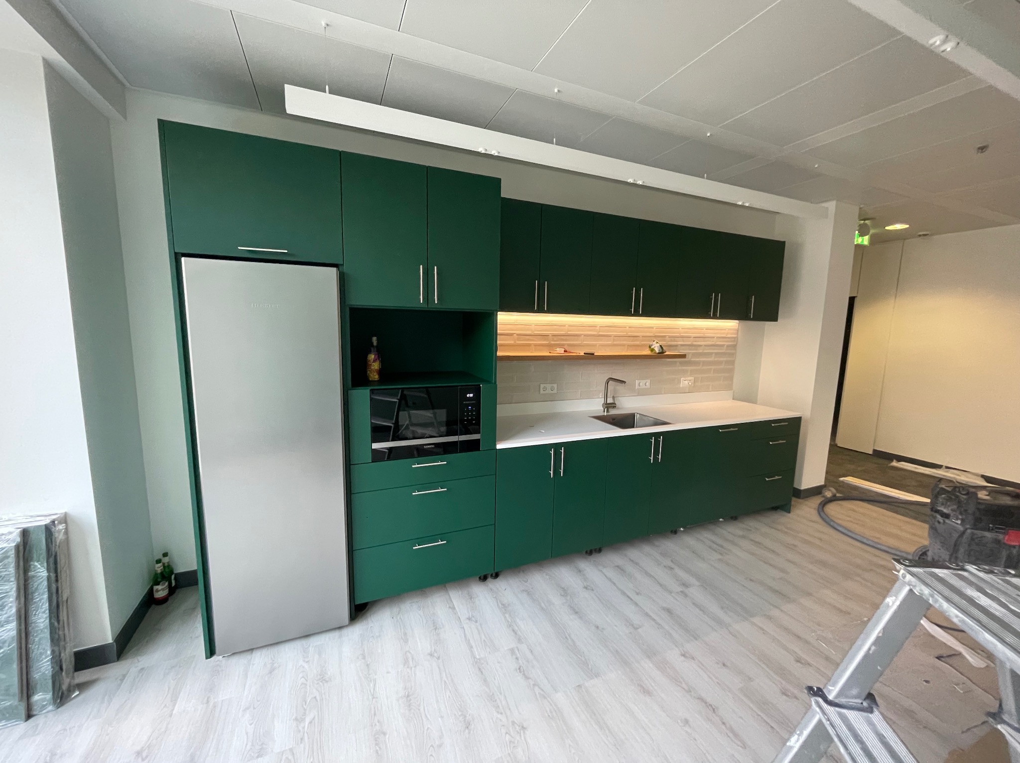 Nowoczesna kuchnia w biurze z zielonymi szafkami, białym blatem i sprzętem AGD, widoczny odkurzacz przemysłowy i drabina w tle.