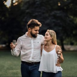 Studio Fotograficzne BARTŁOMIEJ SUCHANEK - Zakochana para w plenerze, on obejmuje ją ramieniem, trzymając kieliszek wina. Ona spogląda na niego z uśmiechem, również z kieliszkiem. Romantyczna sesja na tle zieleni.