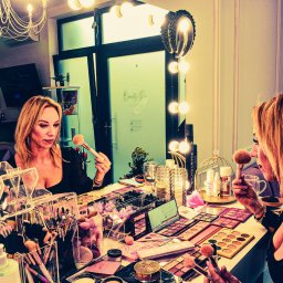 Studio Fotograficzne BARTŁOMIEJ SUCHANEK - Kobieta z blond włosami, wizażystka, aplikuje puder pędzlem przed lustrem z oświetleniem. Na stole palety cieni, pędzle i kosmetyki. Salon w tle.