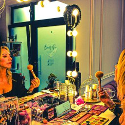 Studio Fotograficzne BARTŁOMIEJ SUCHANEK - Kobieta z blond włosami, wizażystka, trzyma pędzel do makijażu przed lustrem z oświetleniem, na stole palety cieni i akcesoria do makijażu. Studio wizażu.