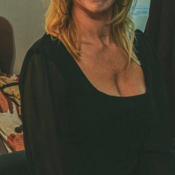 Studio Fotograficzne BARTŁOMIEJ SUCHANEK - Portret uśmiechniętej blondynki w czarnej bluzce z dekoltem, złote kolczyki koła, naturalny makijaż, ciepłe oświetlenie, neutralne tło.