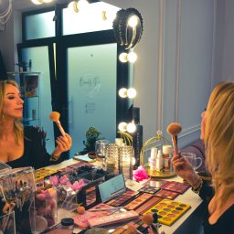 Studio Fotograficzne BARTŁOMIEJ SUCHANEK - Kobieta z blond włosami, w czarnej bluzce, trzyma pędzel do makijażu przed lustrem w salonie kosmetycznym. Na stole palety cieni i akcesoria do makijażu.