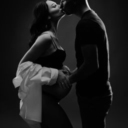 Studio Fotograficzne BARTŁOMIEJ SUCHANEK - Czarno-białe zdjęcie całującej się pary, kobieta w ciąży trzymająca dłoń na brzuchu, w tle ciemne tło, subtelne oświetlenie.