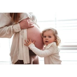 Studio Fotograficzne BARTŁOMIEJ SUCHANEK - Jasne zdjęcie: mała dziewczynka dotyka brzucha ciężarnej mamy, oboje ubrani w beżowe ubrania na tle okna z żaluzjami. Delikatne światło podkreśla bliskość.