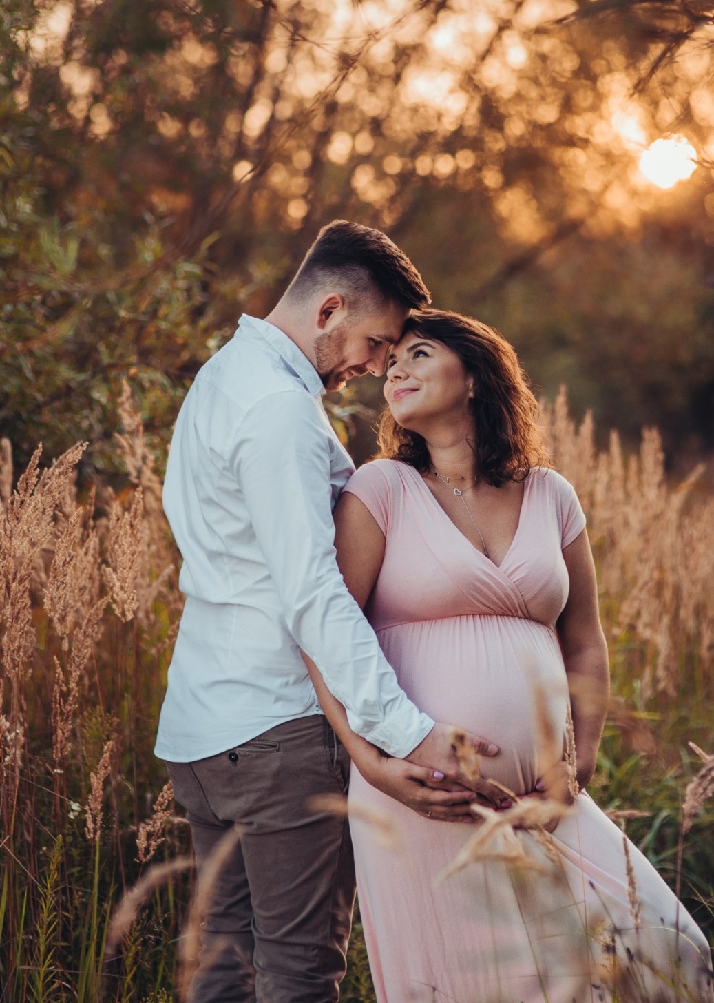 Zakochana para w plenerze: przyszła mama w różowej sukience, czuły dotyk na brzuszku, ciepłe światło zachodzącego słońca. Romantyczna sesja ciążowa.