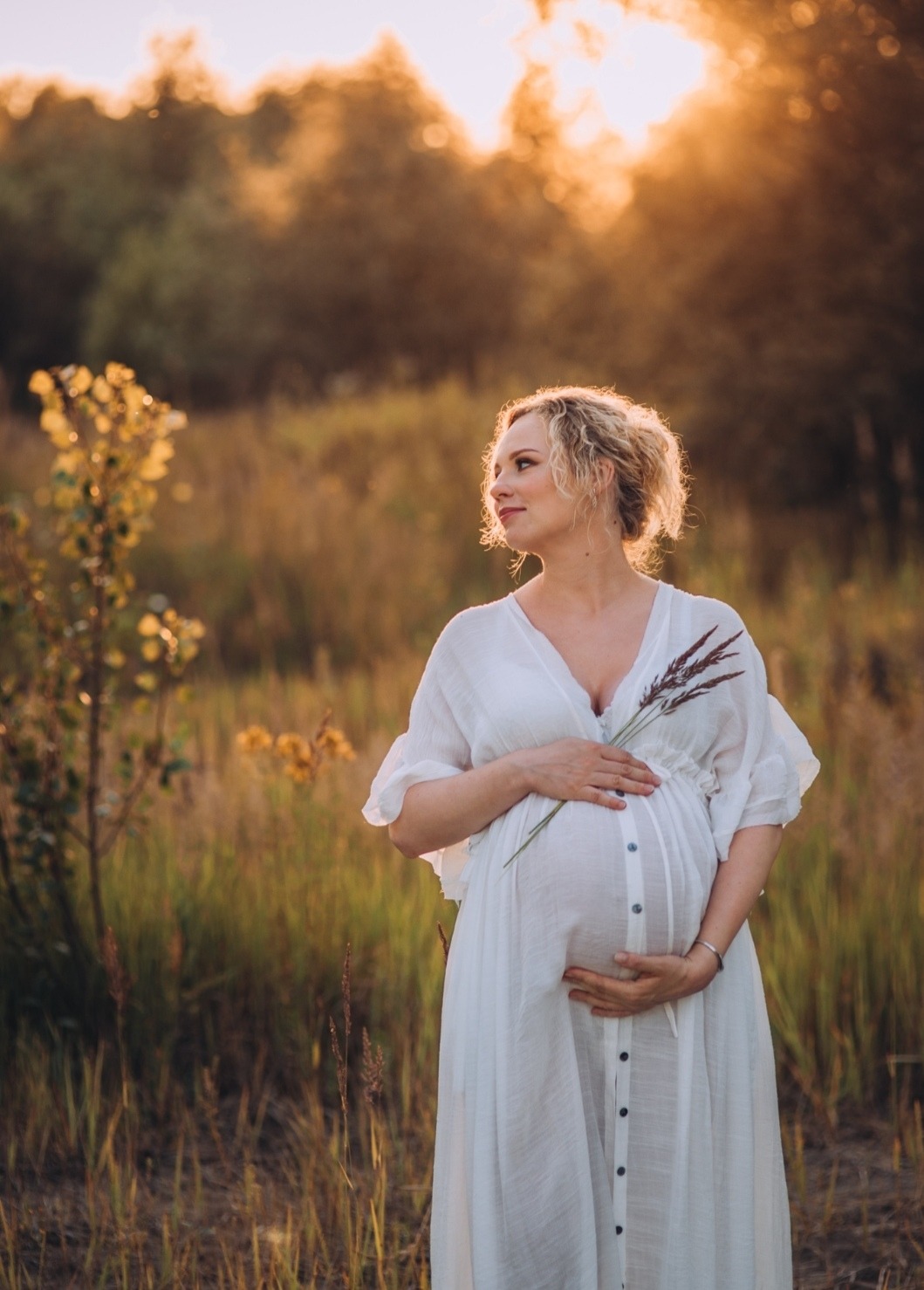 Kobieta w białej sukience, trzymająca źdźbła trawy, z widocznym ciążowym brzuchem, pozuje na łące w ciepłym, zachodzącym słońcu. Delikatne światło podkreśla jej promienny uśmiech.