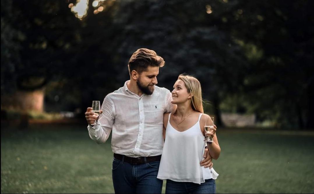 Zakochana para w plenerze, on obejmuje ją ramieniem, trzymając kieliszek wina. Ona spogląda na niego z uśmiechem, również z kieliszkiem. Romantyczna sesja na tle zieleni.