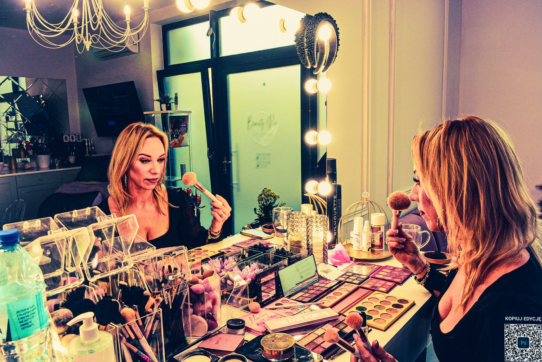 Kobieta z blond włosami, wizażystka, aplikuje puder pędzlem przed lustrem z oświetleniem. Na stole palety cieni, pędzle i kosmetyki. Salon w tle.