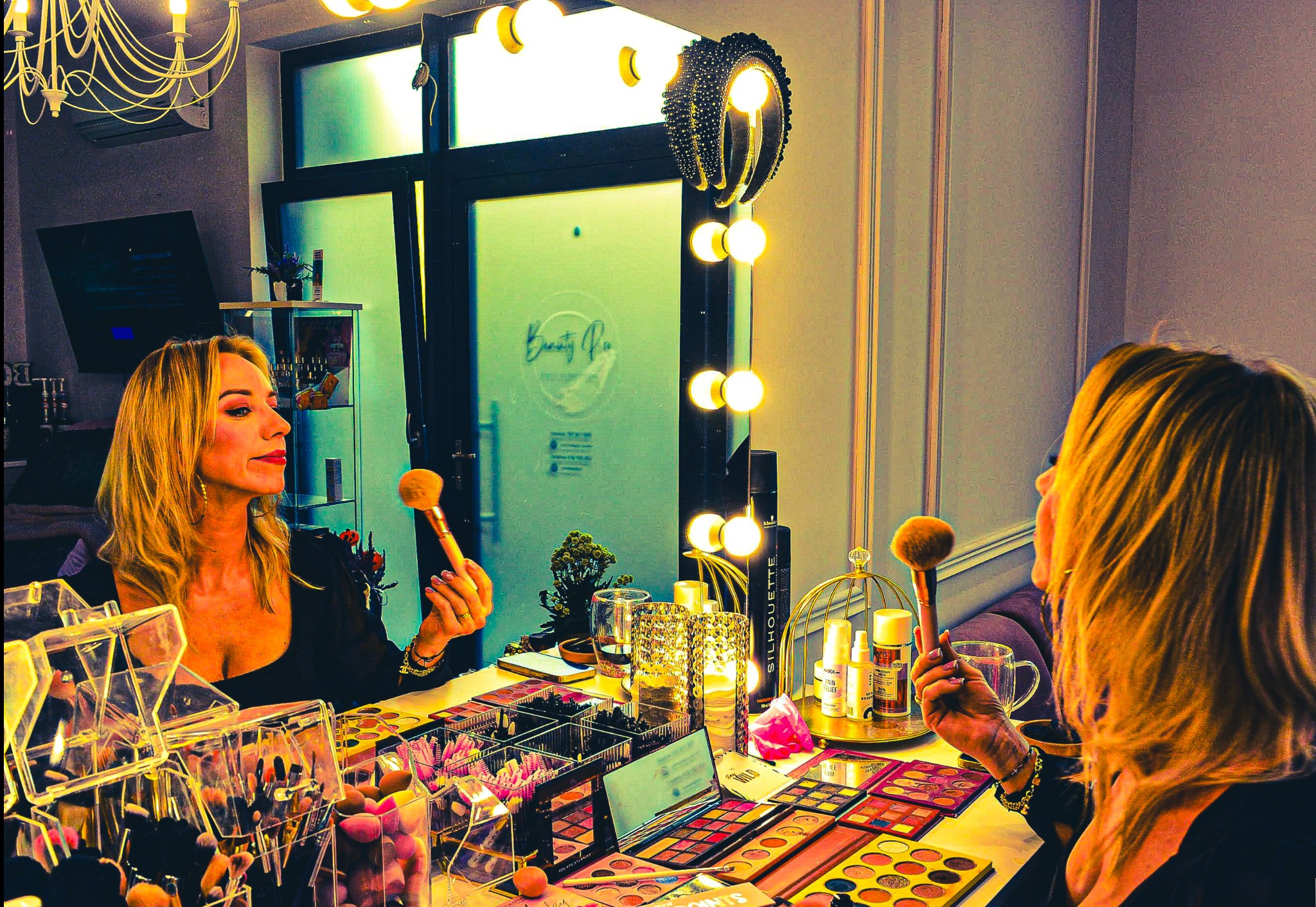 Kobieta z blond włosami, wizażystka, trzyma pędzel do makijażu przed lustrem z oświetleniem, na stole palety cieni i akcesoria do makijażu. Studio wizażu.