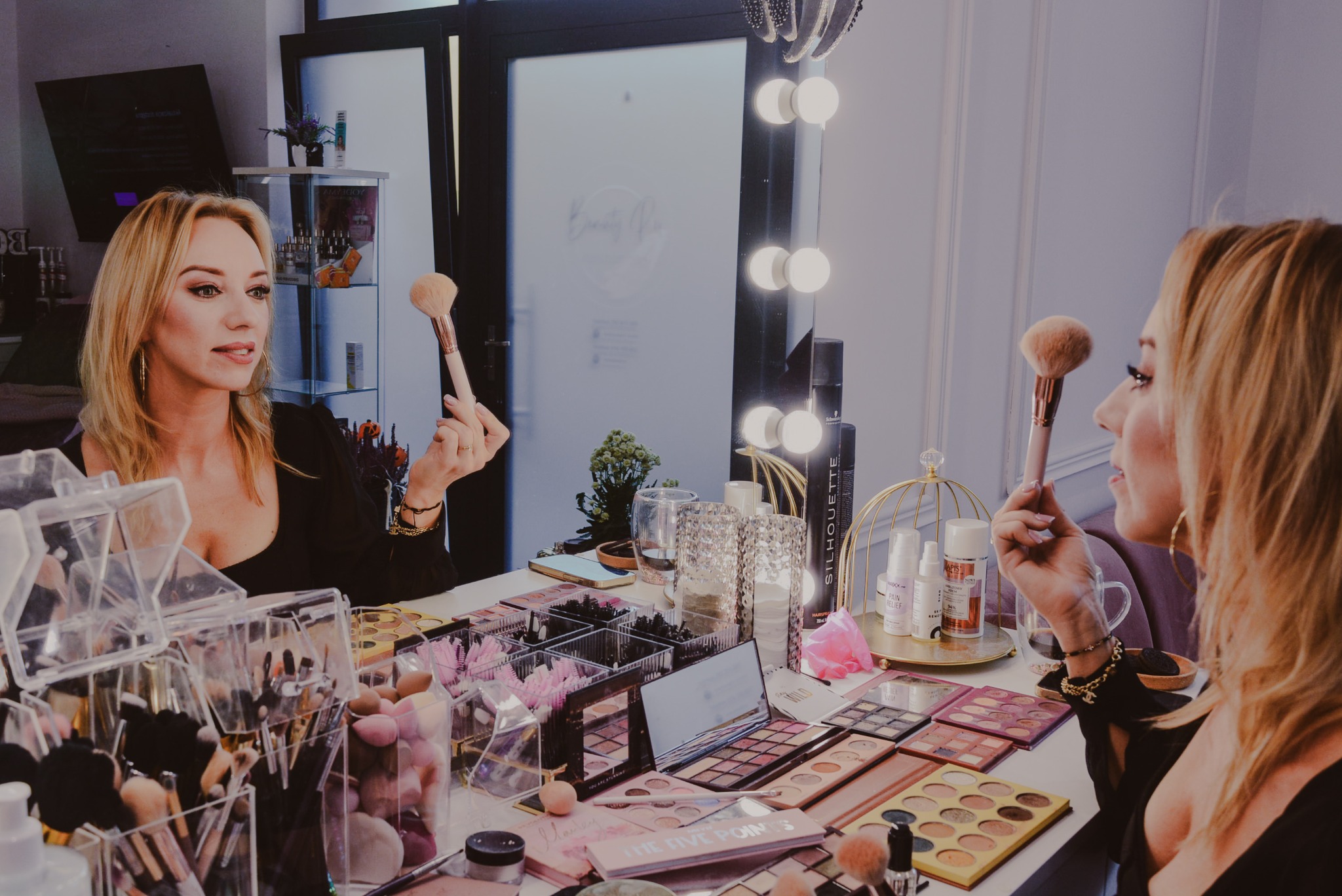 Blond makijażystka z pędzlem w dłoni, odbija się w lustrze z oświetleniem. Na stole palety cieni, pędzle i kosmetyki. Ujęcie z bliska, ciepłe światło.
