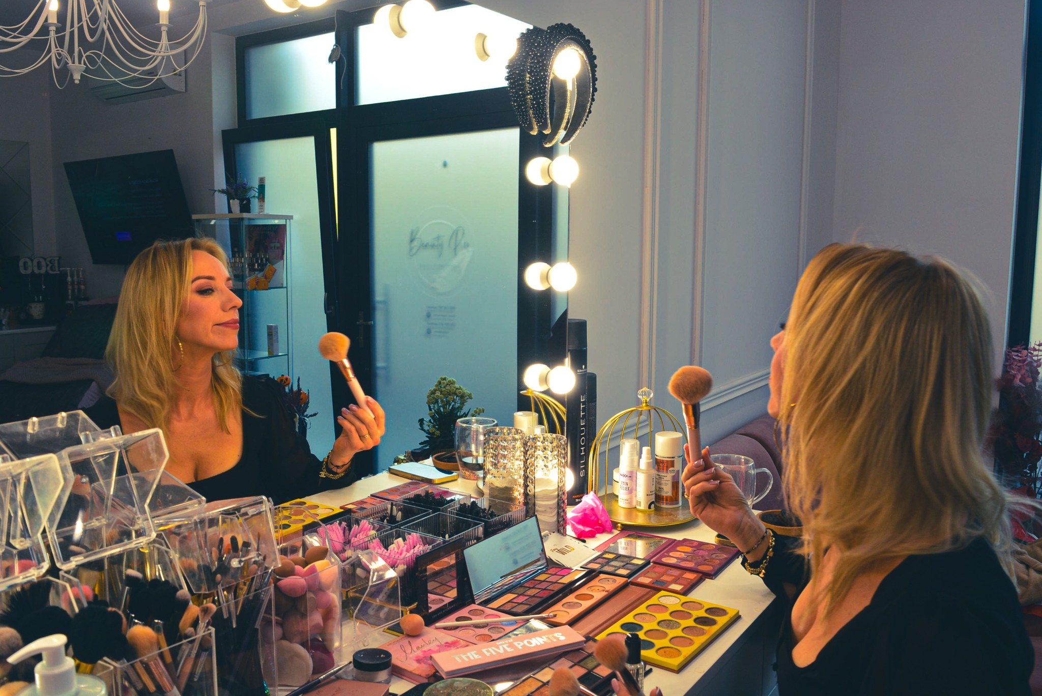 Kobieta z blond włosami, w czarnej bluzce, trzyma pędzel do makijażu przed lustrem w salonie kosmetycznym. Na stole palety cieni i akcesoria do makijażu.