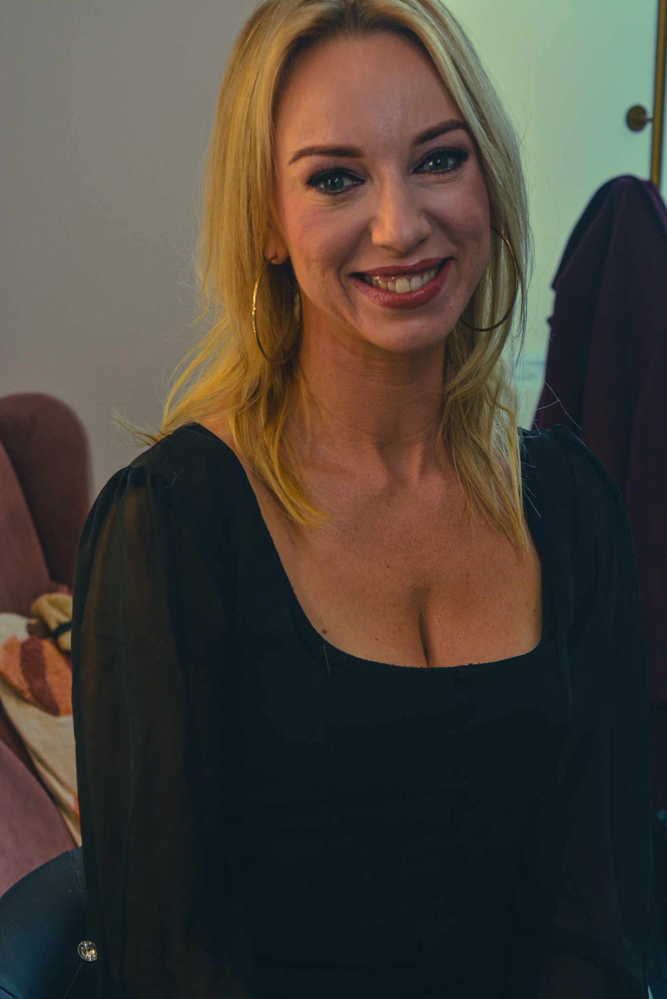 Uśmiechnięta blondynka w czarnej bluzce z dużymi złotymi kolczykami, portret w naturalnym świetle, z delikatnym makijażem i widocznym fragmentem wnętrza.