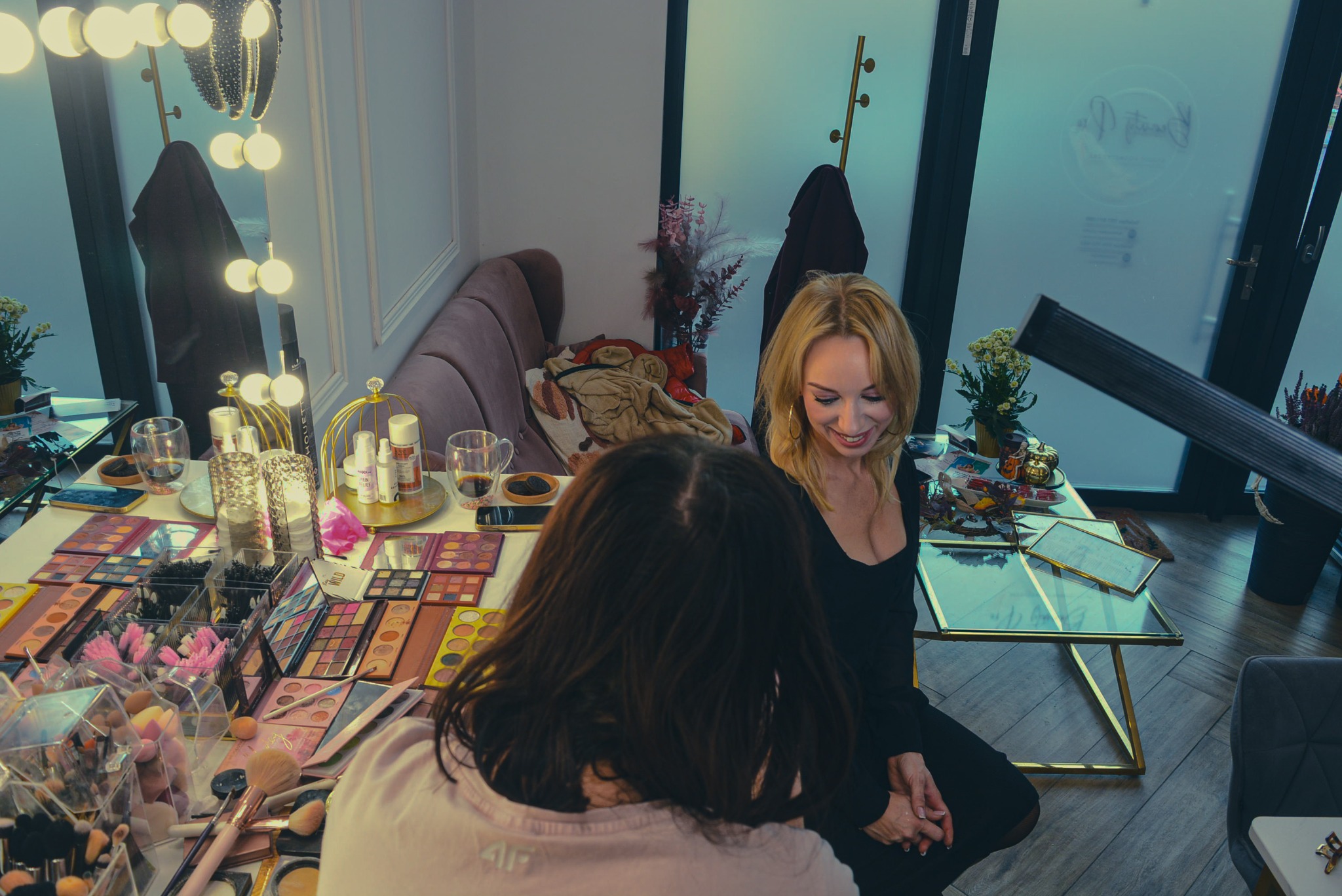 Kobieta z blond włosami uśmiecha się podczas sesji makijażu. Na stole palety cieni, pędzle i kosmetyki. W tle lustro z oświetleniem i sofa.