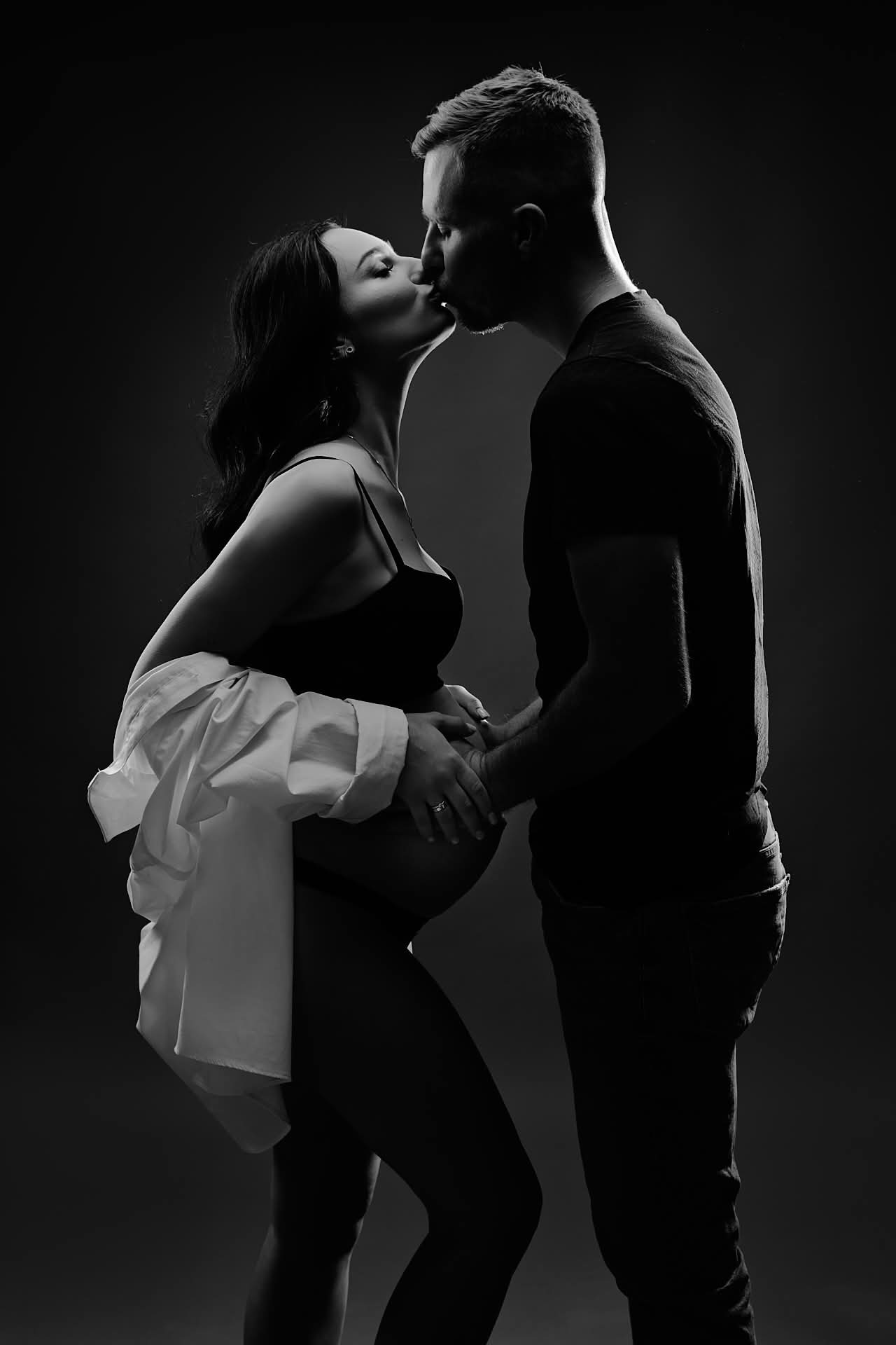 Czarno-białe zdjęcie całującej się pary, kobieta w ciąży trzymająca dłoń na brzuchu, w tle ciemne tło, subtelne oświetlenie.