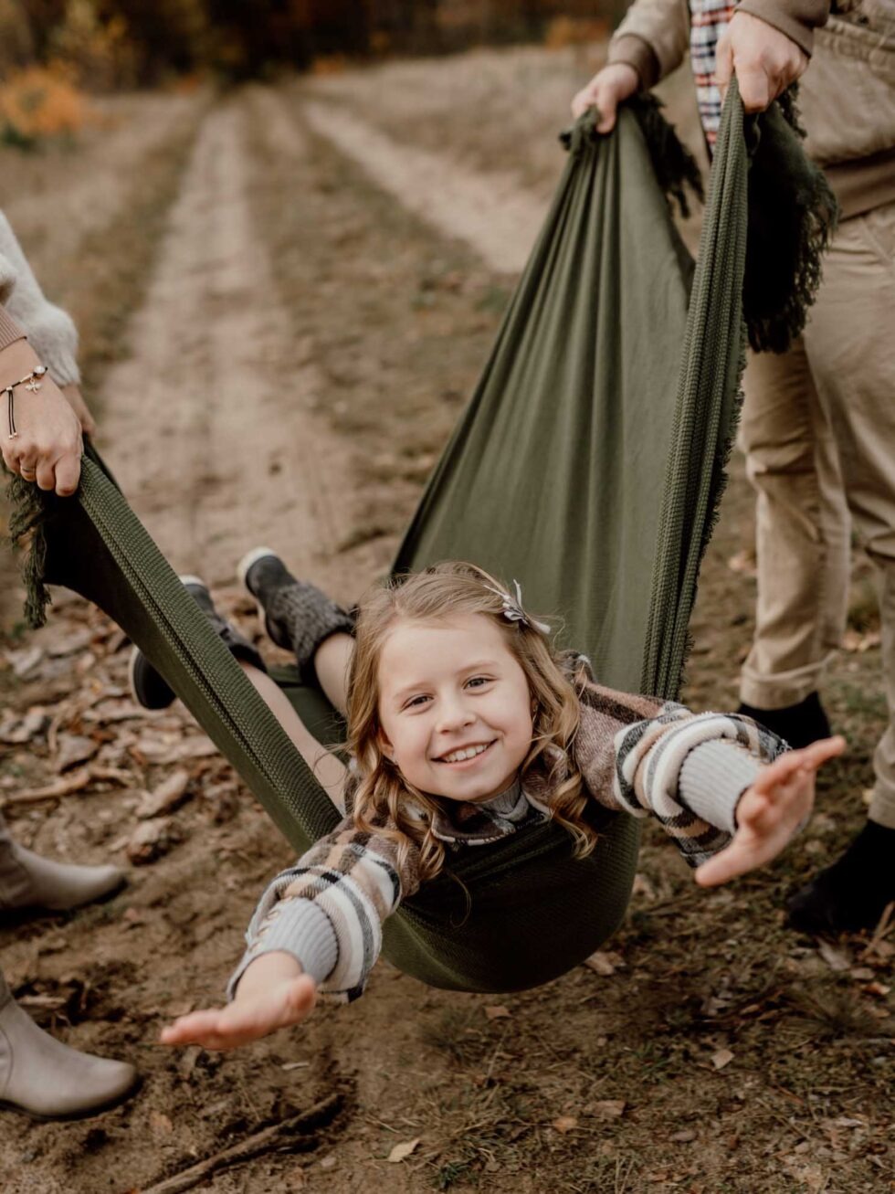 Uśmiechnięta dziewczynka w hamaku z zielonego materiału, trzymanym przez dwie osoby na tle leśnej ścieżki w jesiennej scenerii. Ciepłe, naturalne światło.