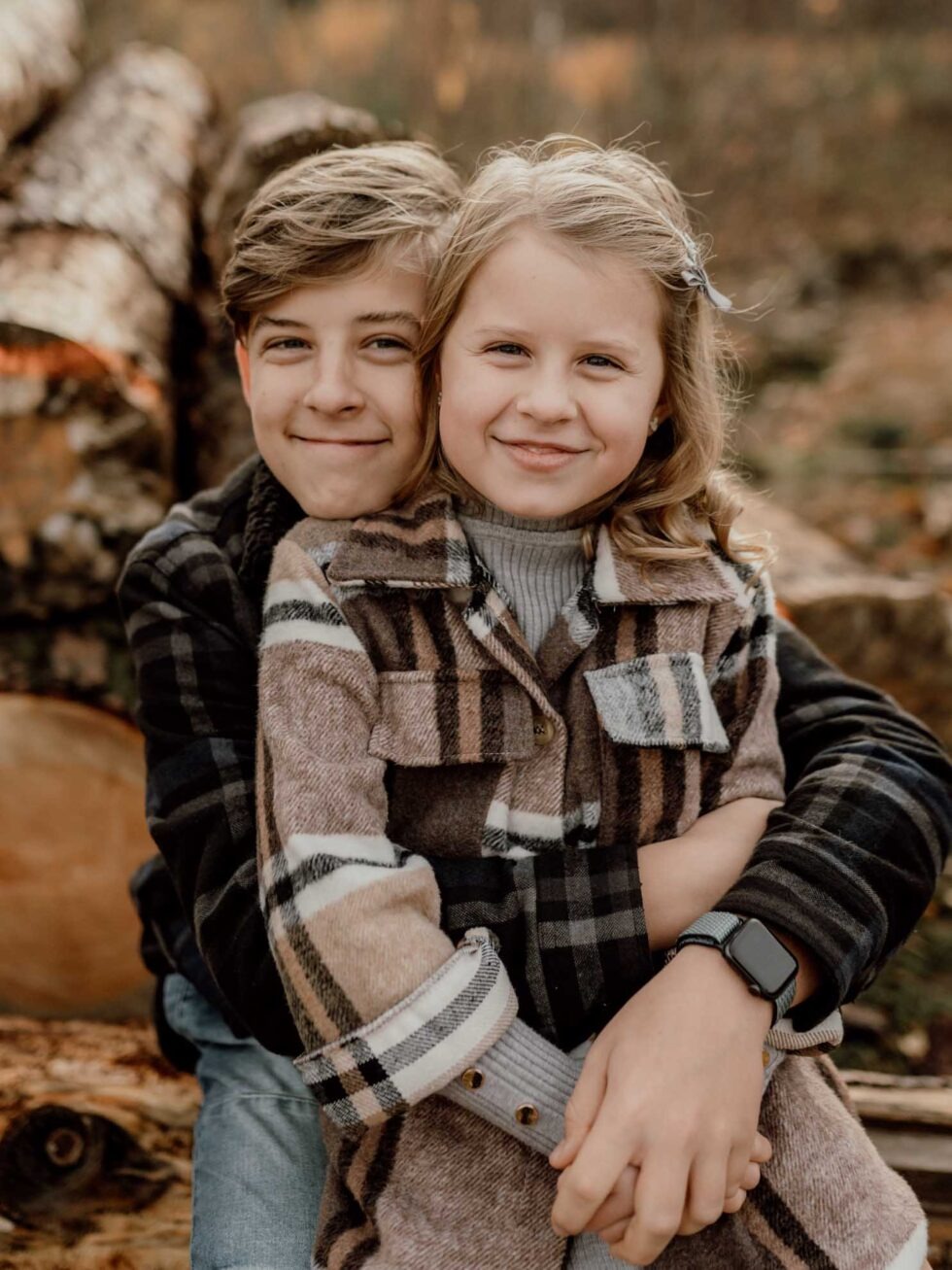 Portret rodzeństwa w jesiennej scenerii: chłopiec obejmuje dziewczynkę w brązowej kraciastej koszuli, oboje uśmiechnięci, w tle stos pni drzew.