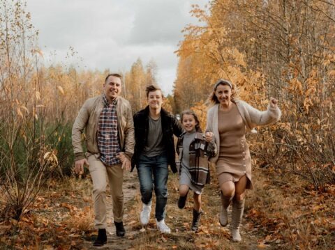 Uśmiechnięta rodzina biegnie ścieżką w jesiennym lesie. Tata w beżowej kurtce, mama w sukience, syn w czarnej kurtce, córka w płaszczu. Ciepłe barwy jesieni.