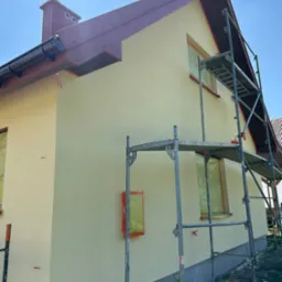 Ocieplona elewacja domu jednorodzinnego w Nowym Sączu, z zabezpieczonymi oknami folią i rusztowaniem w trakcie prac wykończeniowych.