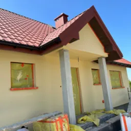 Prace malarskie przy elewacji domu jednorodzinnego, okna i drzwi zabezpieczone folią, widoczny fragment rusztowania i pracownik w trakcie pracy.