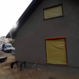 Boczny widok domu w trakcie prac elewacyjnych; okna zabezpieczone folią, świeżo nałożony szary tynk, widoczne przewody i elementy instalacji na zewnątrz.