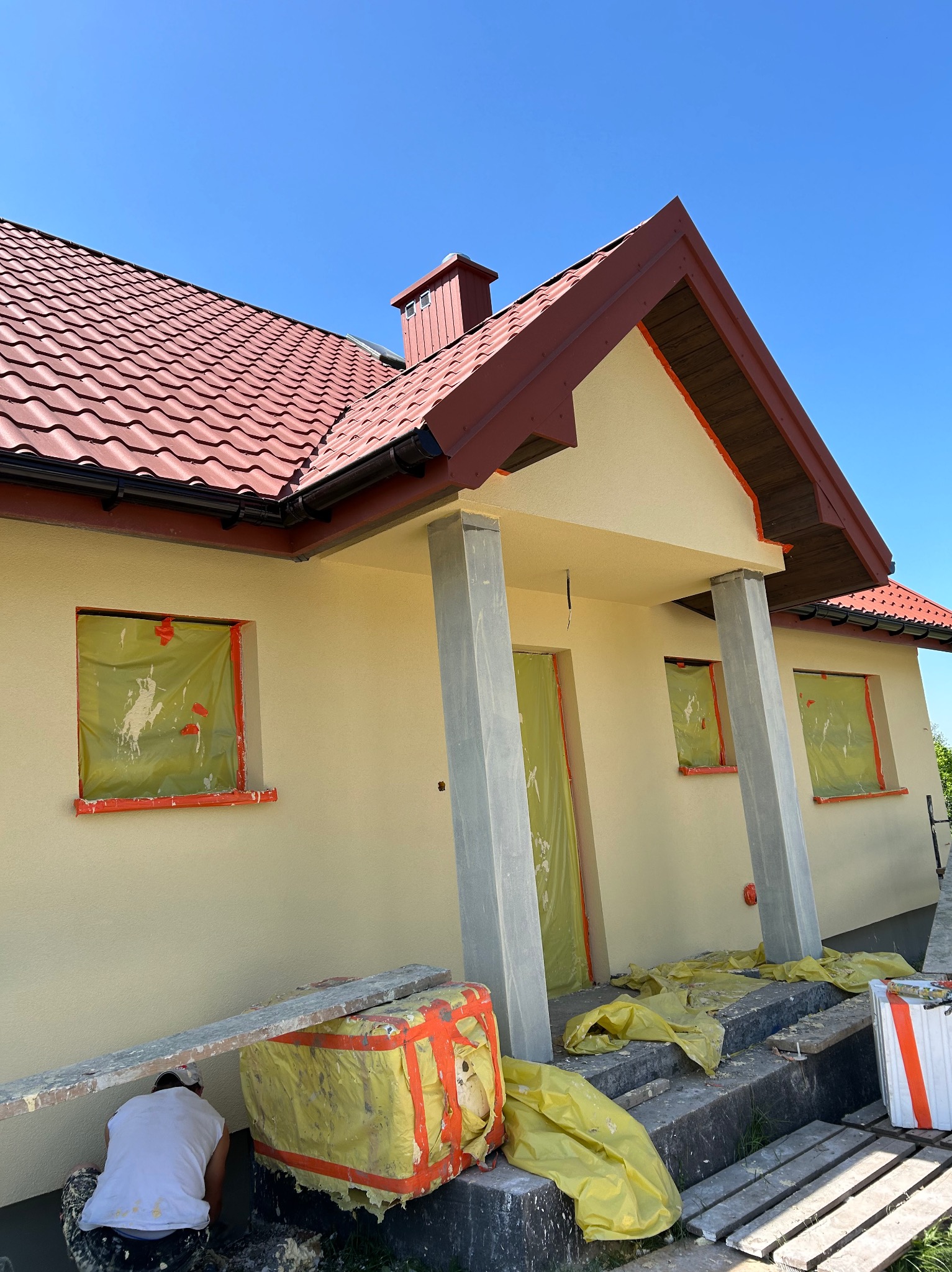 Prace malarskie przy elewacji domu jednorodzinnego, okna i drzwi zabezpieczone folią, widoczny fragment rusztowania i pracownik w trakcie pracy.