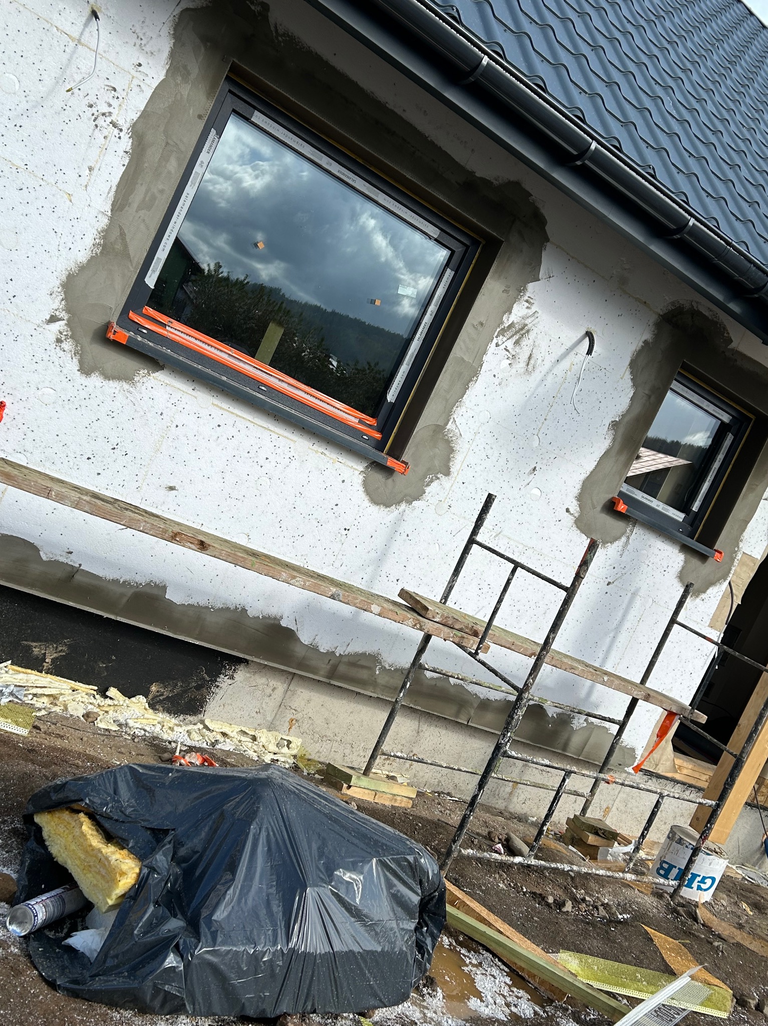 Fragment ściany budynku w trakcie ocieplania: styropian, siatka i zaprawa wokół okien, widoczne rusztowanie i odpady budowlane w worku.