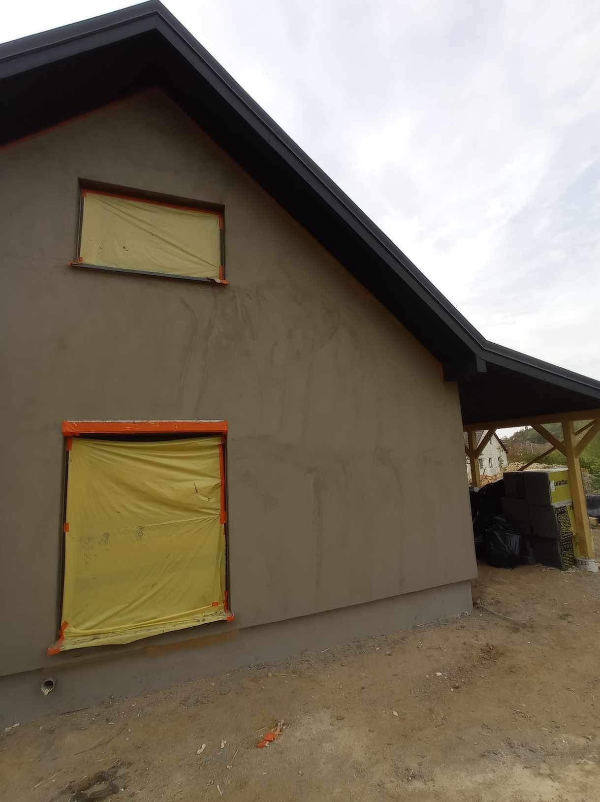 Elewacja domu w trakcie prac: szara ściana z zabezpieczonymi oknami folią i pomarańczową taśmą malarską, widoczne ślady świeżego tynku, czarne wykończenie dachu, materiały budowlane pod zadaszeniem...