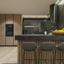 AG Design & Estate - Nowoczesna kuchnia z drewnianymi szafkami, czarnym blatem i trzema szarymi krzesłami barowymi, oświetlona paskami LED, z owocami w plecionym koszu na blacie.