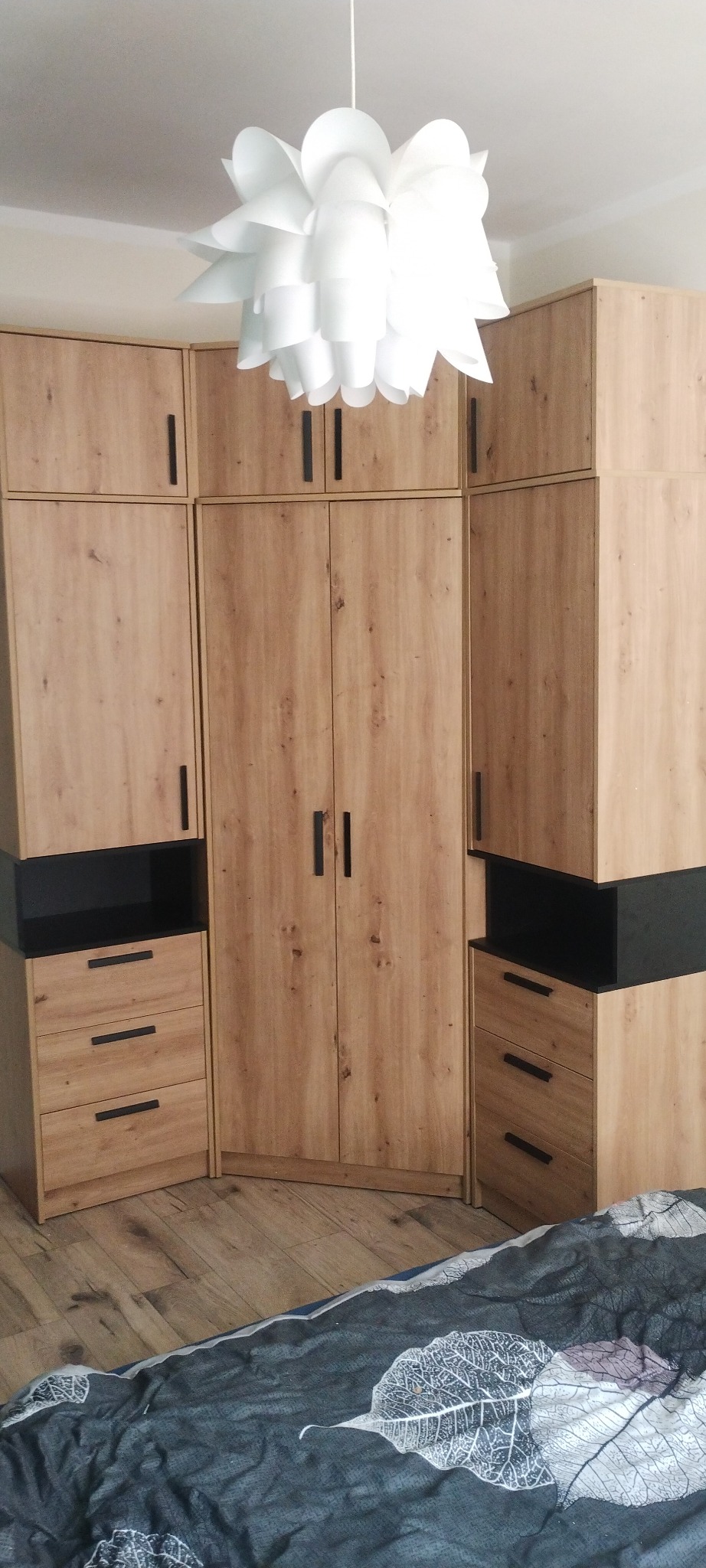 Komplet szaf z drewnopodobną okleiną i czarnymi elementami, tworzący zabudowę w rogu pokoju, pod nowoczesną białą lampą.