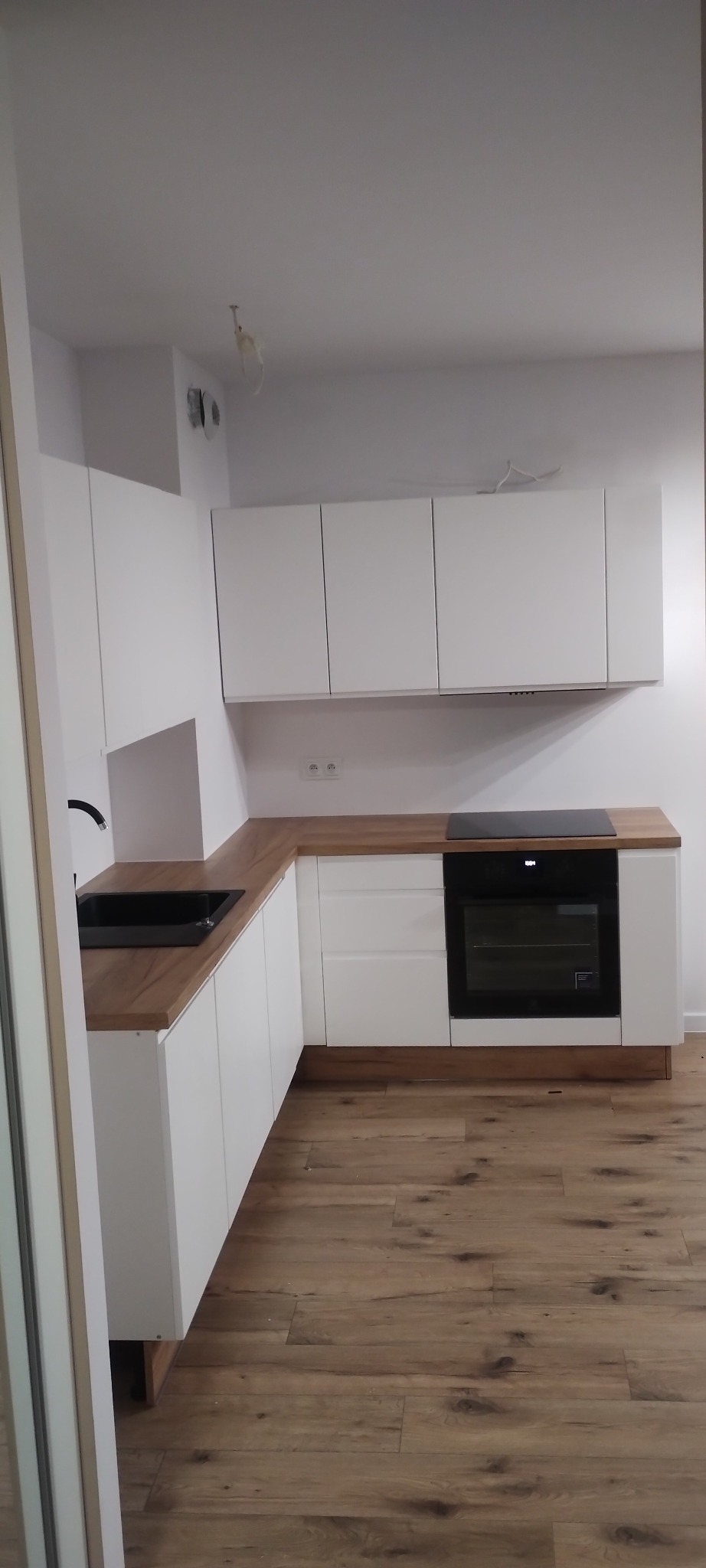 Nowoczesna, jasna kuchnia z białymi szafkami i drewnianym blatem, czarnym zlewem i płytą indukcyjną, na podłodze panele imitujące drewno.