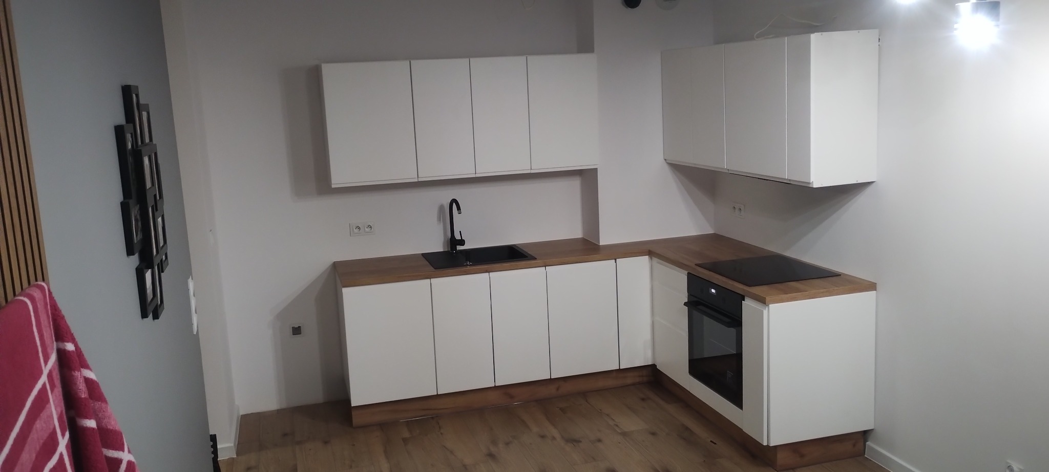 Nowoczesna, minimalistyczna kuchnia z białymi szafkami i drewnianym blatem, czarnym zlewem i płytą indukcyjną, oświetlona punktowymi lampami sufitowymi.