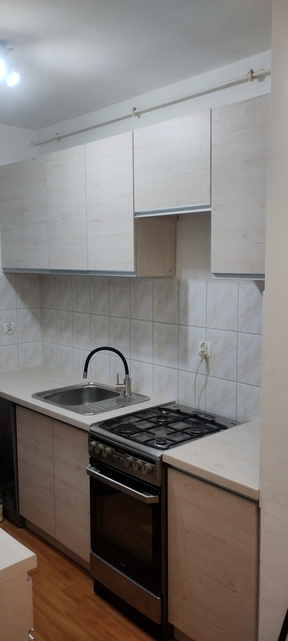 Nowoczesna kuchnia z jasnymi szafkami, czarną armaturą i płytą gazową, widok na świeżo wyremontowane wnętrze z drewnianą podłogą.