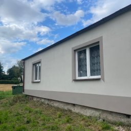 Michał Szymczuk - Świeżo pomalowana elewacja budynku z dwoma oknami z białymi ramami i koronkowymi firankami, widoczny cokół w ciemniejszym odcieniu szarości, na tle błękitnego nieba z chmurami i zieleni trawy.