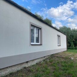 Michał Szymczuk - Świeżo pomalowana elewacja budynku z dwoma oknami, widoczna szara podmurówka i fragment trawnika w pochmurny dzień.