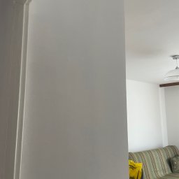 Michał Szymczuk - Świeżo pomalowana, jasnoszara ściana z widocznym włącznikiem światła, fragmentem futryny i częściowo widocznym pokojem z kanapą w tle.