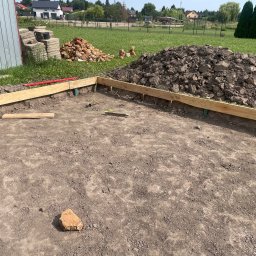 Michał Szymczuk - Przygotowanie terenu pod fundament: drewniany szalunek, poziomica, cegły i pryzma ziemi na trawiastym tle.