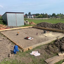 Michał Szymczuk - Przygotowanie fundamentów pod dom murowany: drewniany szalunek, narzędzia budowlane i sterta cegieł na trawiastym terenie.