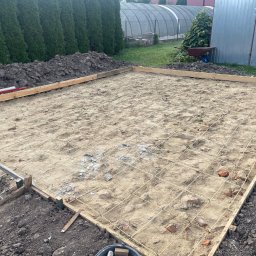 Michał Szymczuk - Przygotowany teren pod wylewkę fundamentów z drewnianym szalunkiem, siatką zbrojeniową i fragmentami cegieł, widok z góry na tle zieleni i szarej blaszanej ściany.